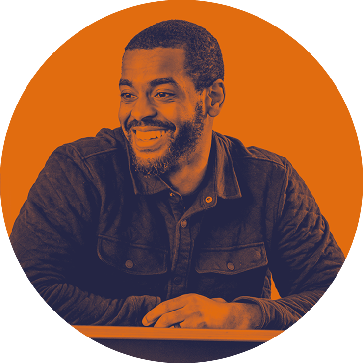 Smiling man in a dark shirt sitting at a table with an orange circular background - Mix Talent